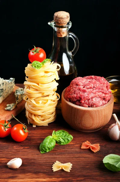 Italian food concept. Ingredients for cooking pasta, such as pasta, olive oil, ground beef, cheese and basil leaves on a brown wooden board. Black background. Place for writing text
