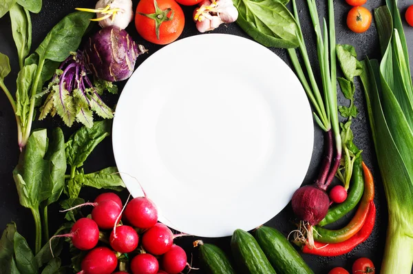 The white plate surrounded by vegetables and herbs, such as cherry tomatoes, radishes, green onions, spinach, garlic and hot peppers. Vegetable background. Vegan concept