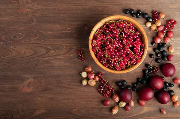 Summer berry background (wallpaper). Plate with berries red currant on a brown wooden board. Near a scattering of other fruits and berries, such as gooseberries, black currants and plums. Space for text. Vegan concept