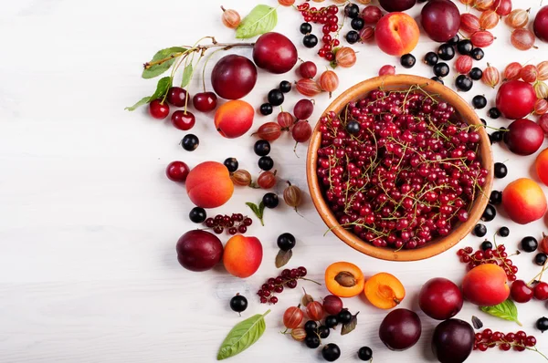 Summer berry background (wallpaper). Plate with berries red currant on a white wooden board. Near a scattering of other fruits and berries, such as gooseberries, black currants, apricots and plums. Space for text. Vegan concept