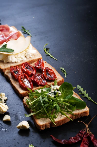 The concept of Italian food. Sandwiches with various fillings, such as arugula, dried tomatoes, pear, blue cheese and ham on dark background