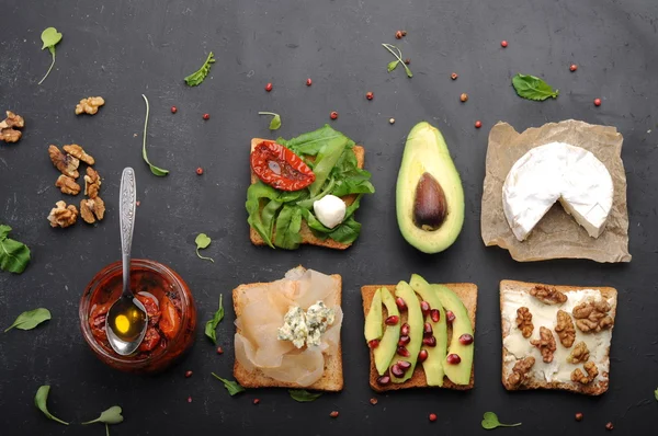 Sandwiches with a variety of toppings such as arugula, dried tomatoes, soft cheese, blue cheese, nuts and avocados on a dark background. The concept of healthy vegetarian food.