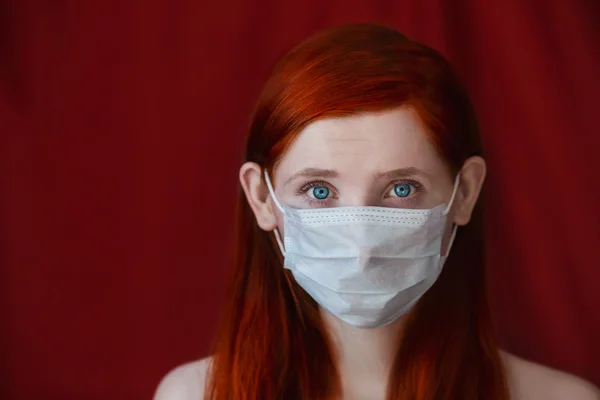Red-haired girl with a medical mask on a red background, woman doctor