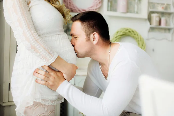 Happy man kissing belly of his lovely pregnant wife standing in bedroom.