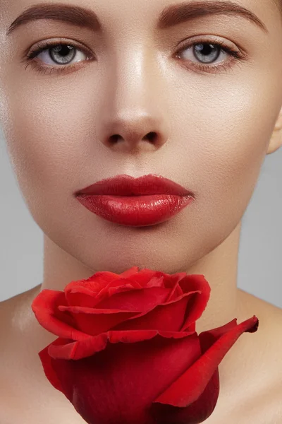 Close-up beautiful young woman with bright lipgloss makeup. Perfect clean skin, sexy red lip make-up. Beautiful valentine visage with red rose flower. Romantic and sexy look for Valentines day