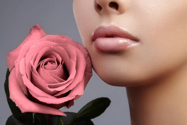 Close-up beautiful female lips with bright lipgloss makeup. Perfect clean skin, light fresh lip make-up. Beautiful spa portrait with tender pink rose flower. Spa and cosmetics