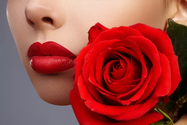 Close-up beautiful female lips with bright lipgloss makeup. Perfect clean skin, sexy red lip make-up. Beautiful visage portrait with red rose flower. Visage and cosmetics