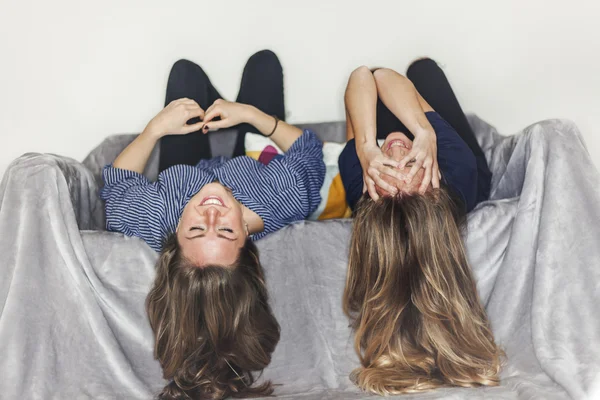 Two girls laying upside down on a grey sofa at home smiling