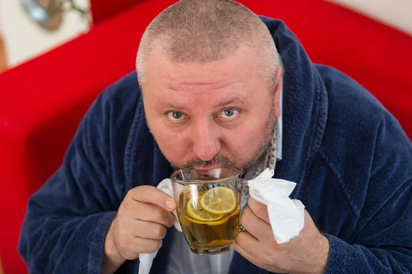 Ill man blowing his nose with paper napkin and drinking tea at home