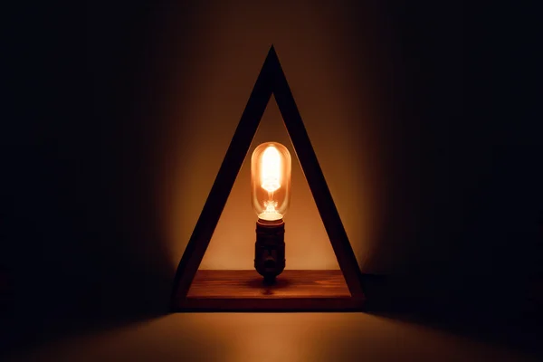 Classic Edison old glowing light bulb with filament in loft lamp standing in wooden triangl as a candle. Abstract background. eco led illuminator at night