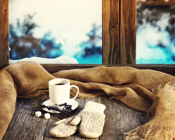 White cup of coffee or tea, lavender flowers, mittens and natural gunny cloth located on stylized wooden window sill. Winter concept of comfort and relaxation. Photo with retro filter effect.