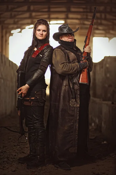 Man with rifle in a leather garment and woman in raider costume with crossbow inside a concrete shelter.