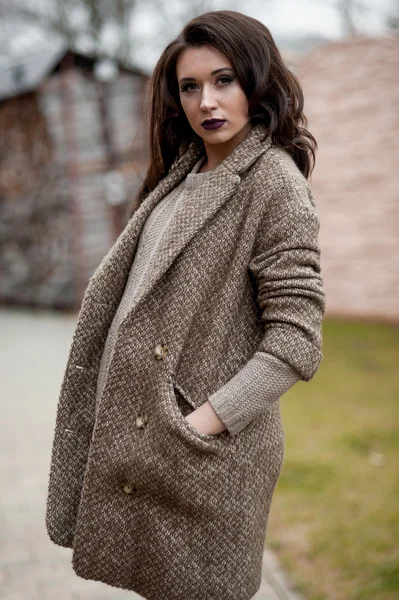 Spring in the country style. Full length portrait of young woman in white knitted sweater and furry hat standing in the front of rustic wood wall in the yard near house, wearing sweater casual.