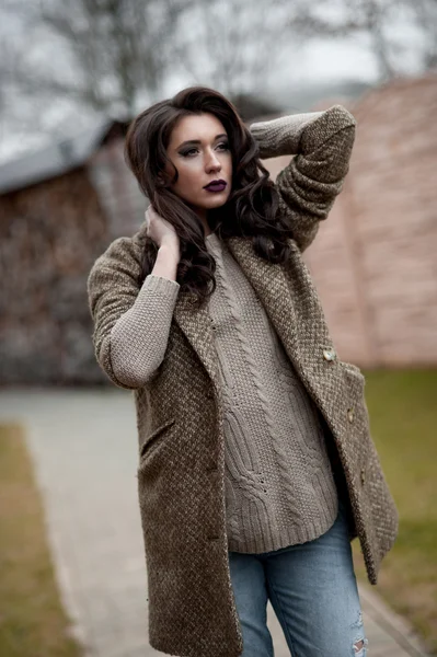 Spring in the country style. Full length portrait of young woman in white knitted sweater and furry hat standing in the front of rustic wood wall in the yard near house, wearing sweater casual.