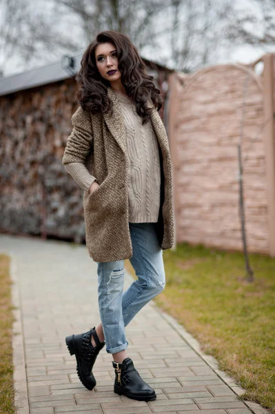 Spring in the country style. Full length portrait of young woman in white knitted sweater and furry hat standing in the front of rustic wood wall in the yard near house, wearing sweater casual.