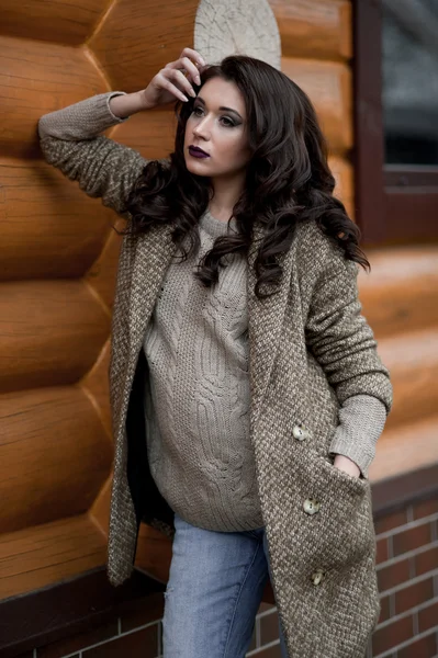 Spring in the country style. Full length portrait of young woman in white knitted sweater and furry hat standing in the front of rustic wood wall in the yard near house, wearing sweater casual.