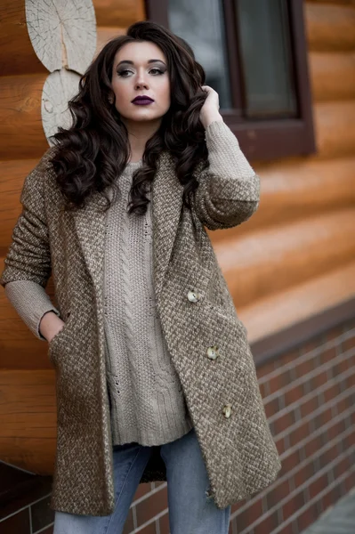 Spring in the country style. Full length portrait of young woman in white knitted sweater and furry hat standing in the front of rustic wood wall in the yard near house, wearing sweater casual.