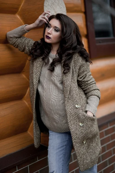 Spring in the country style. Full length portrait of young woman in white knitted sweater and furry hat standing in the front of rustic wood wall in the yard near house, wearing sweater casual.