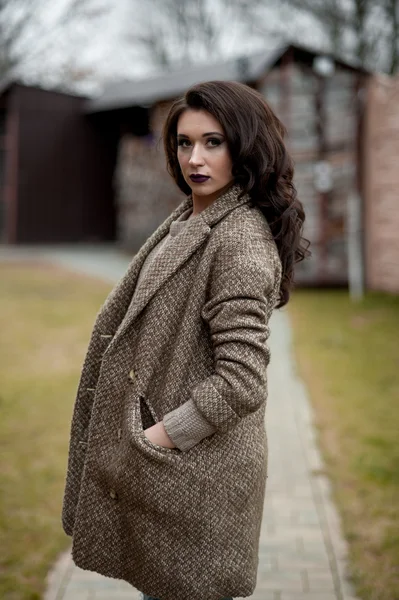 Spring in the country style. Full length portrait of young woman in white knitted sweater and furry hat standing in the front of rustic wood wall in the yard near house, wearing sweater casual.