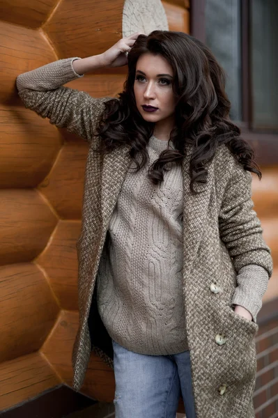 Spring in the country style. Full length portrait of young woman in white knitted sweater and furry hat standing in the front of rustic wood wall in the yard near house, wearing sweater casual.