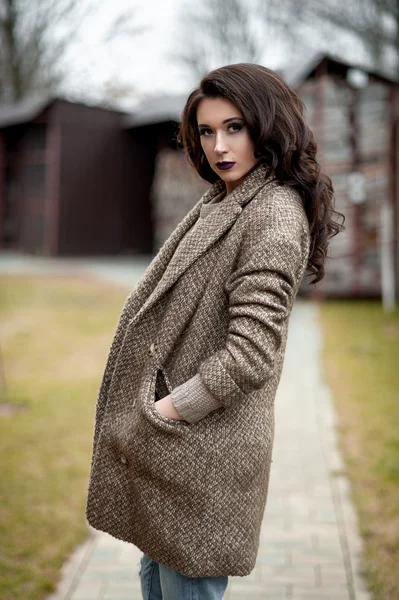 Spring in the country style. Full length portrait of young woman in white knitted sweater and furry hat standing in the front of rustic wood wall in the yard near house, wearing sweater casual.