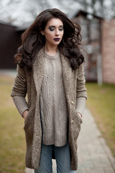 Spring in the country style. Full length portrait of young woman in white knitted sweater and furry hat standing in the front of rustic wood wall in the yard near house, wearing sweater casual.