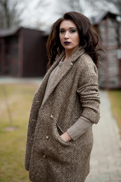 Spring in the country style. Full length portrait of young woman in white knitted sweater and furry hat standing in the front of rustic wood wall in the yard near house, wearing sweater casual.