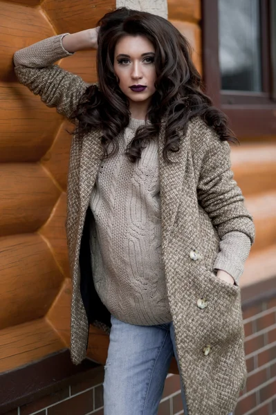 Spring in the country style. Full length portrait of young woman in white knitted sweater and furry hat standing in the front of rustic wood wall in the yard near house, wearing sweater casual.