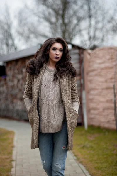 Spring in the country style. Full length portrait of young woman in white knitted sweater and furry hat standing in the front of rustic wood wall in the yard near house, wearing sweater casual.