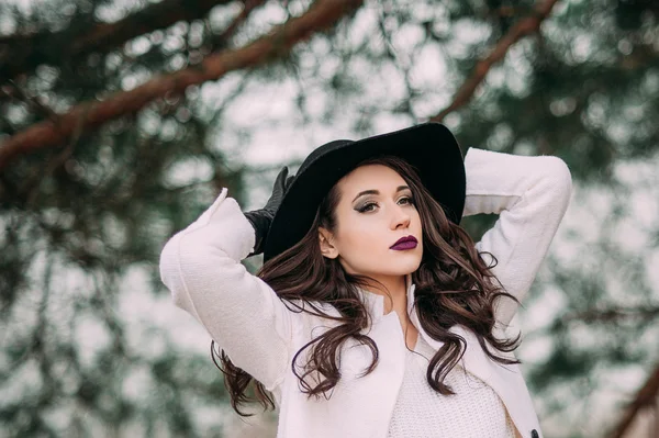 Beautiful fashionable woman in a black hat and white coat posing outdoors
