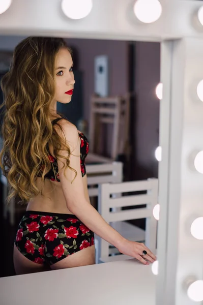 Young beautiful woman applying her make up face with brush, looking in a mirror, sitting on chair at dressing room with vintage mirror dark room, wearing red lingerie