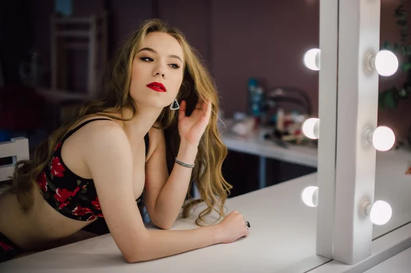 Young beautiful woman applying her make up face with brush, looking in a mirror, sitting on chair at dressing room with vintage mirror dark room, wearing red lingerie