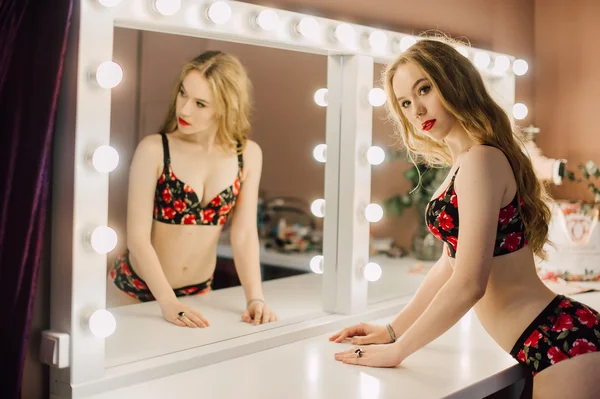 Young beautiful woman applying her make up face with brush, looking in a mirror, sitting on chair at dressing room with vintage mirror dark room, wearing red lingerie