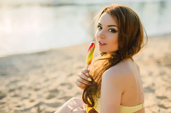 Summer vacation, travel, tourism, junk food and people concept - young woman in sunglasses eating colorful ice cream on resort beach background, wearing yellow swimsuit