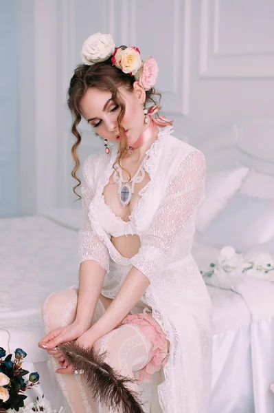 Beautiful young sexy woman sitting on white bed, wearing white lace dress, hair decorated with flowers. Perfect makeup. Beauty fashion. Eyelashes. Studio retouched shot.