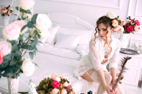 Beautiful young sexy woman sitting on white bed, wearing white lace dress, hair decorated with flowers. Perfect makeup. Beauty fashion. Eyelashes. Studio retouched shot.