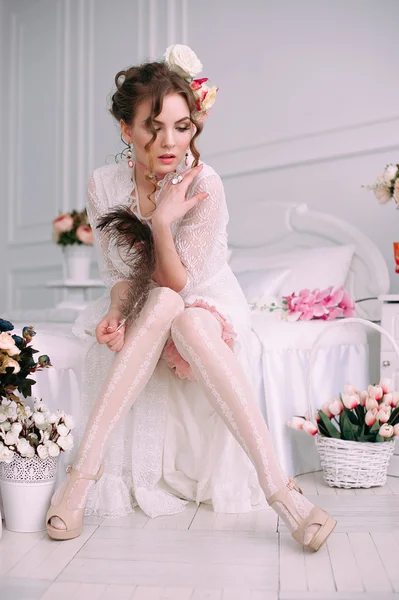 Beautiful young sexy woman sitting on white bed, wearing white lace dress, hair decorated with flowers. Perfect makeup. Beauty fashion. Eyelashes. Studio retouched shot.