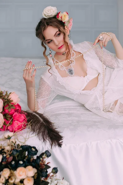 Beautiful young sexy woman sitting on white bed, wearing white lace dress, hair decorated with flowers. Perfect makeup. Beauty fashion. Eyelashes. Studio retouched shot.