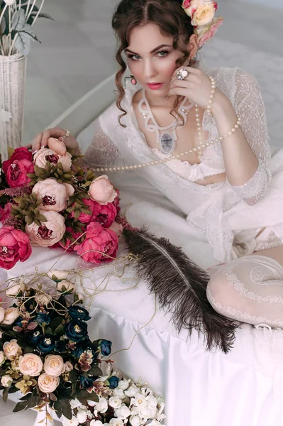 Beautiful young sexy woman sitting on white bed, wearing white lace dress, hair decorated with flowers. Perfect makeup. Beauty fashion. Eyelashes. Studio retouched shot.