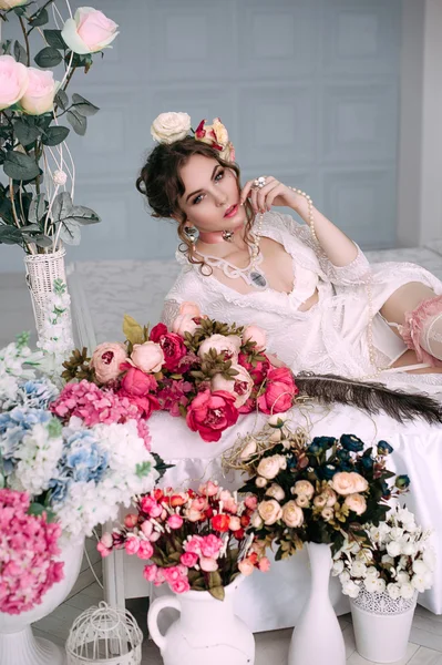 Beautiful young sexy woman sitting on white bed, wearing white lace dress, hair decorated with flowers. Perfect makeup. Beauty fashion. Eyelashes. Studio retouched shot.