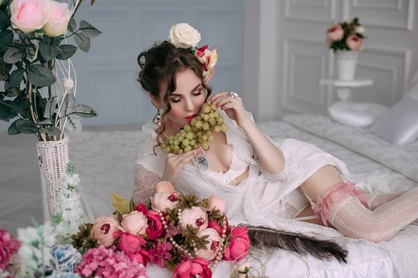 Beautiful young sexy woman sitting on white bed and eating grapes, wearing white lace dress, room decorated with flowers. Perfect makeup. Beauty fashion. Eyelashes. Studio retouched shot.