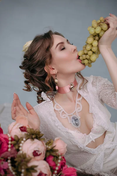 Beautiful young sexy woman sitting on white bed and eating grapes, wearing white lace dress, room decorated with flowers. Perfect makeup. Beauty fashion. Eyelashes. Studio retouched shot.