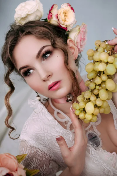 Beautiful young sexy woman sitting on white bed and eating grapes, wearing white lace dress, room decorated with flowers. Perfect makeup. Beauty fashion. Eyelashes. Studio retouched shot.