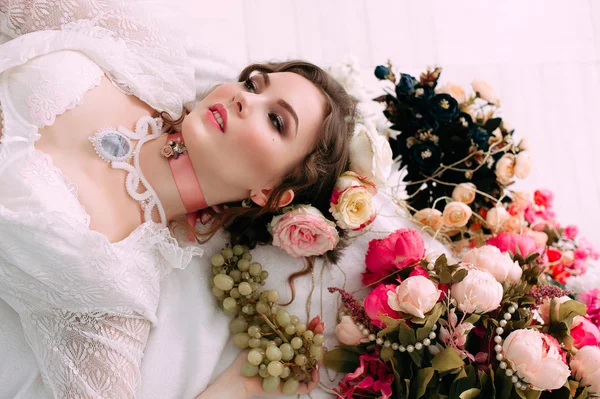 Beautiful young sexy woman sitting on white bed and eating grapes, wearing white lace dress, room decorated with flowers. Perfect makeup. Beauty fashion. Eyelashes. Studio retouched shot.