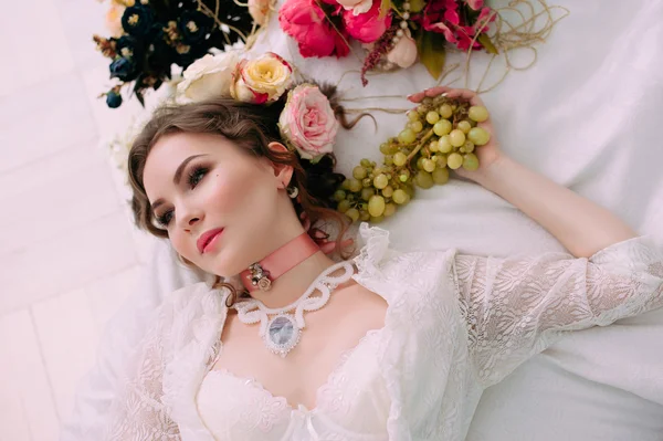 Beautiful young sexy woman sitting on white bed and eating grapes, wearing white lace dress, room decorated with flowers. Perfect makeup. Beauty fashion. Eyelashes. Studio retouched shot.