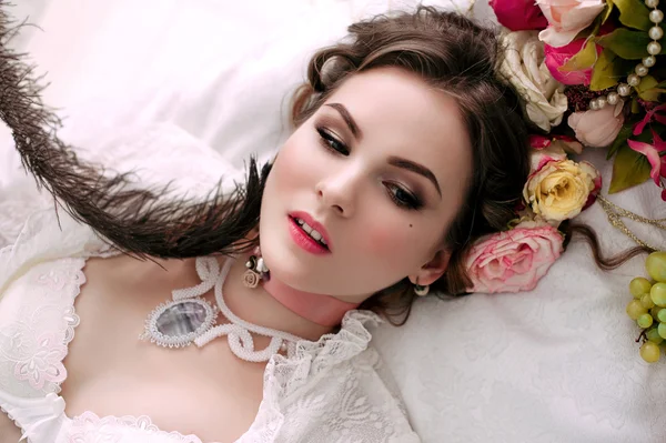 Beautiful young sexy woman sitting on white bed and eating grapes, wearing white lace dress, room decorated with flowers. Perfect makeup. Beauty fashion. Eyelashes. Studio retouched shot.