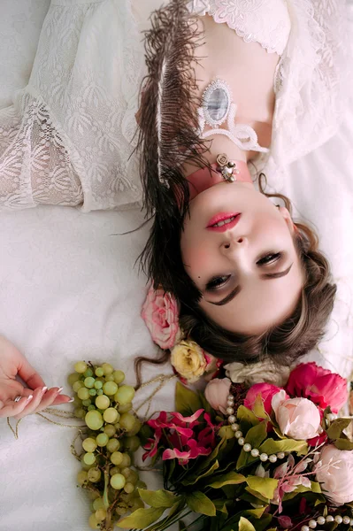 Beautiful young sexy woman sitting on white bed and eating grapes, wearing white lace dress, room decorated with flowers. Perfect makeup. Beauty fashion. Eyelashes. Studio retouched shot.