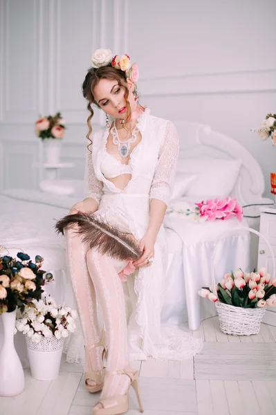 Beautiful young sexy woman sitting on white bed, wearing white lace dress, hair decorated with flowers. Perfect makeup. Beauty fashion. Eyelashes. Studio retouched shot.