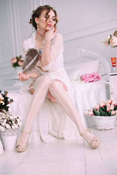 Beautiful young sexy woman sitting on white bed, wearing white lace dress, hair decorated with flowers. Perfect makeup. Beauty fashion. Eyelashes. Studio retouched shot.