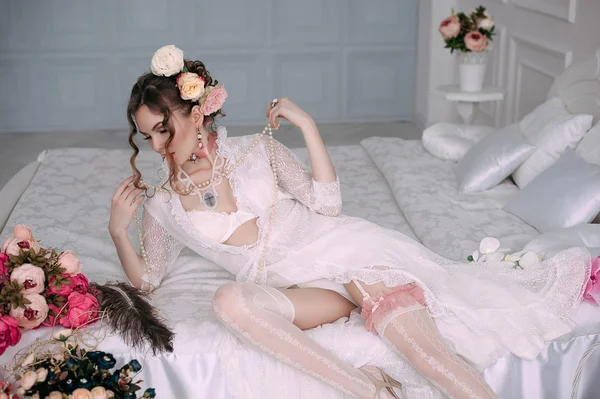 Beautiful young sexy woman sitting on white bed, wearing white lace dress, hair decorated with flowers. Perfect makeup. Beauty fashion. Eyelashes. Studio retouched shot.